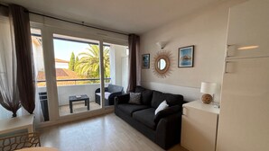 A modern living room with a sofa, a small table, a refrigerator, and a balcony with a view of palm trees. - Charming studio - 4 guests, 2 mins to beach (Urrugne)
