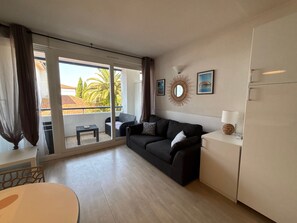 Uma sala de estar moderna com sofá, uma mesinha, geladeira e varanda com vista para palmeiras.