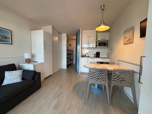 A compact living space with a kitchenette, a dining area with a table and chairs, a sofa with cushions, and a hanging light fixture.