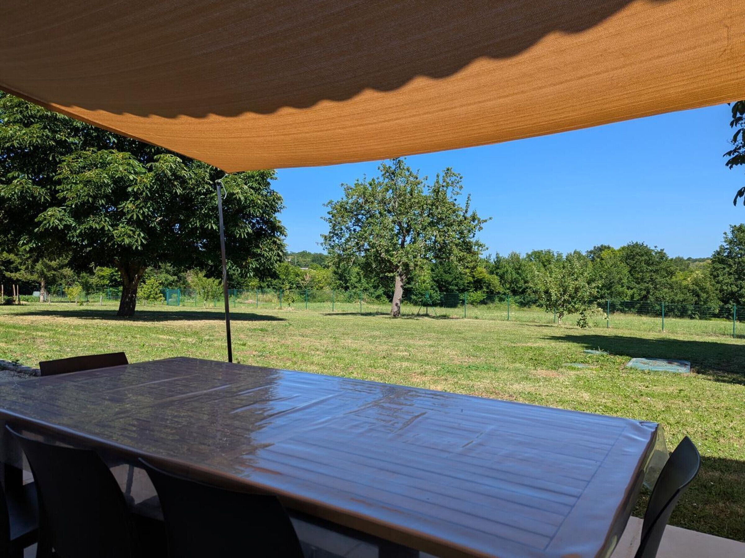 Mesa y sillas de madera bajo una vela de sombra en un espacio exterior con árboles y cielo despejado.