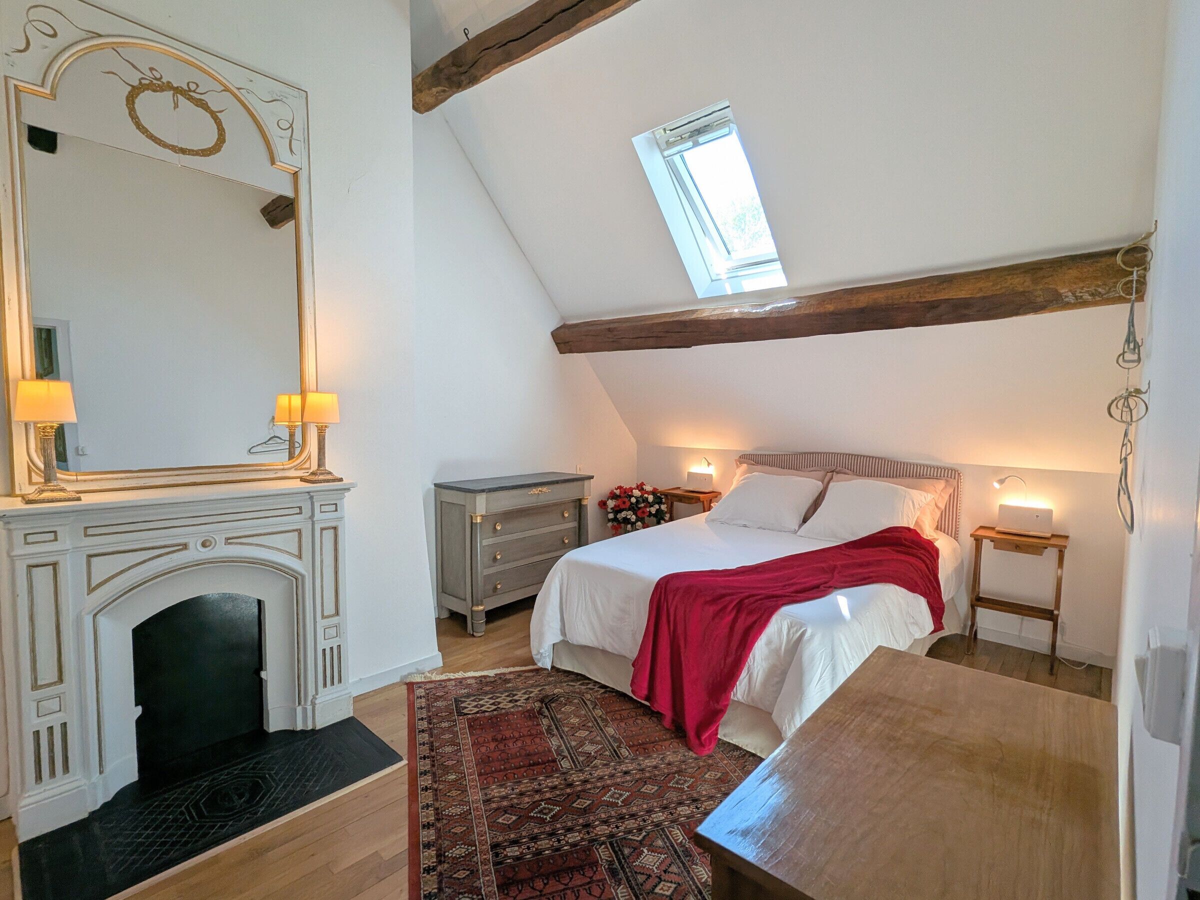 Un dormitorio con cama, cómoda, espejo, chimenea, tragaluz y viga de techo de madera.