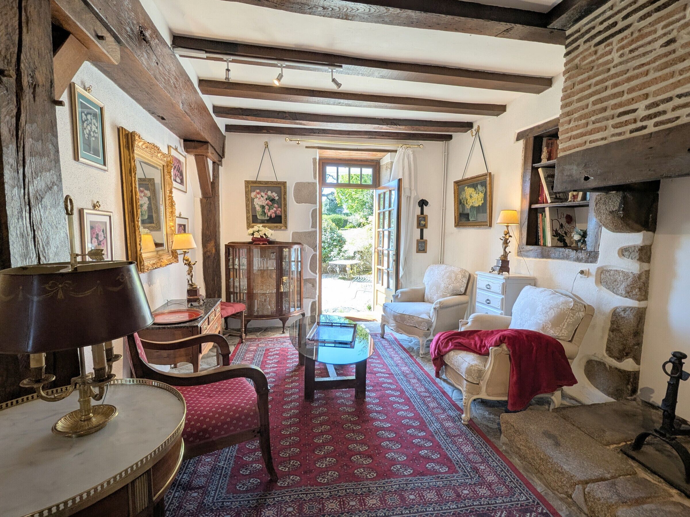 A traditional room with exposed wooden beams, a stone wall, a red patterned rug, and a view through a window.