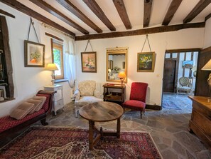 Une pièce chaleureuse avec un sol en pierre, des poutres en bois, un fauteuil rouge, une table ronde en bois et un canapé agrémenté de coussins à motifs.