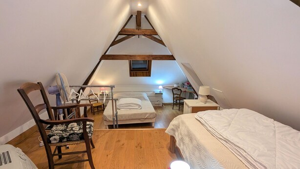 Um quarto de sótão acolhedor, com teto inclinado, vigas de madeira, uma cama, uma cadeira, uma escrivaninha e um abajur.