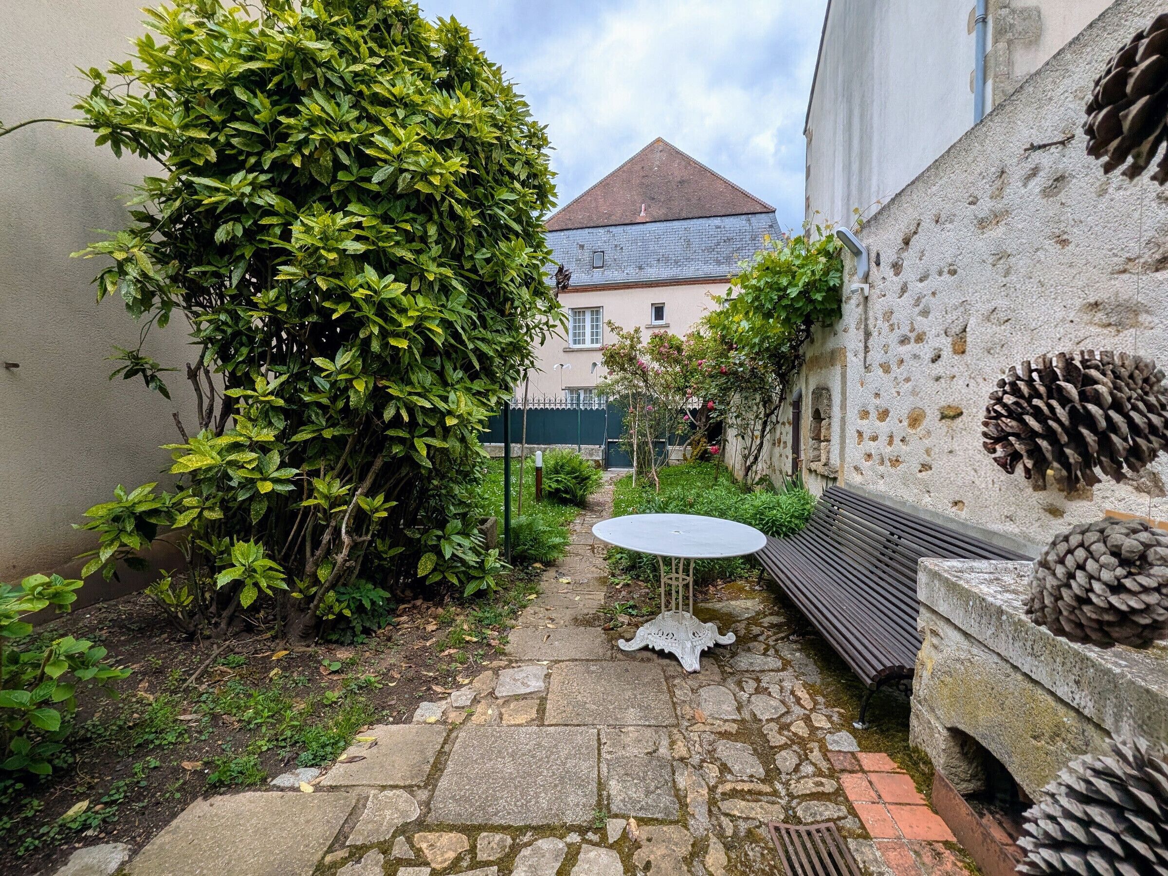 A stone pathway leading to a small round table and bench, surrounded by greenery and a building with a sloped roof.