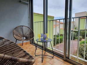 Ein Balkon mit Korbmöbeln, einem kleinen Tisch und zwei Gläsern, mit Blick auf eine Wohngegend.