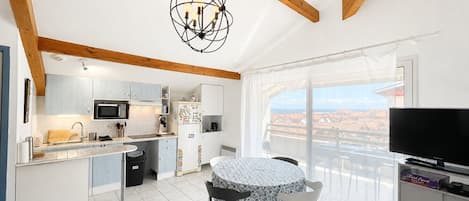 Una cocina moderna con gabinetes blancos, un microondas y un refrigerador. Hay un comedor con una mesa y sillas, y un balcón con vistas.