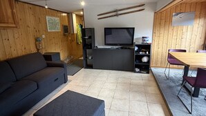 A living room with a sofa, ottoman, TV, wooden walls, and a dining area with chairs. - Studio in the heart of the station - pets welcome (Le Lioran)