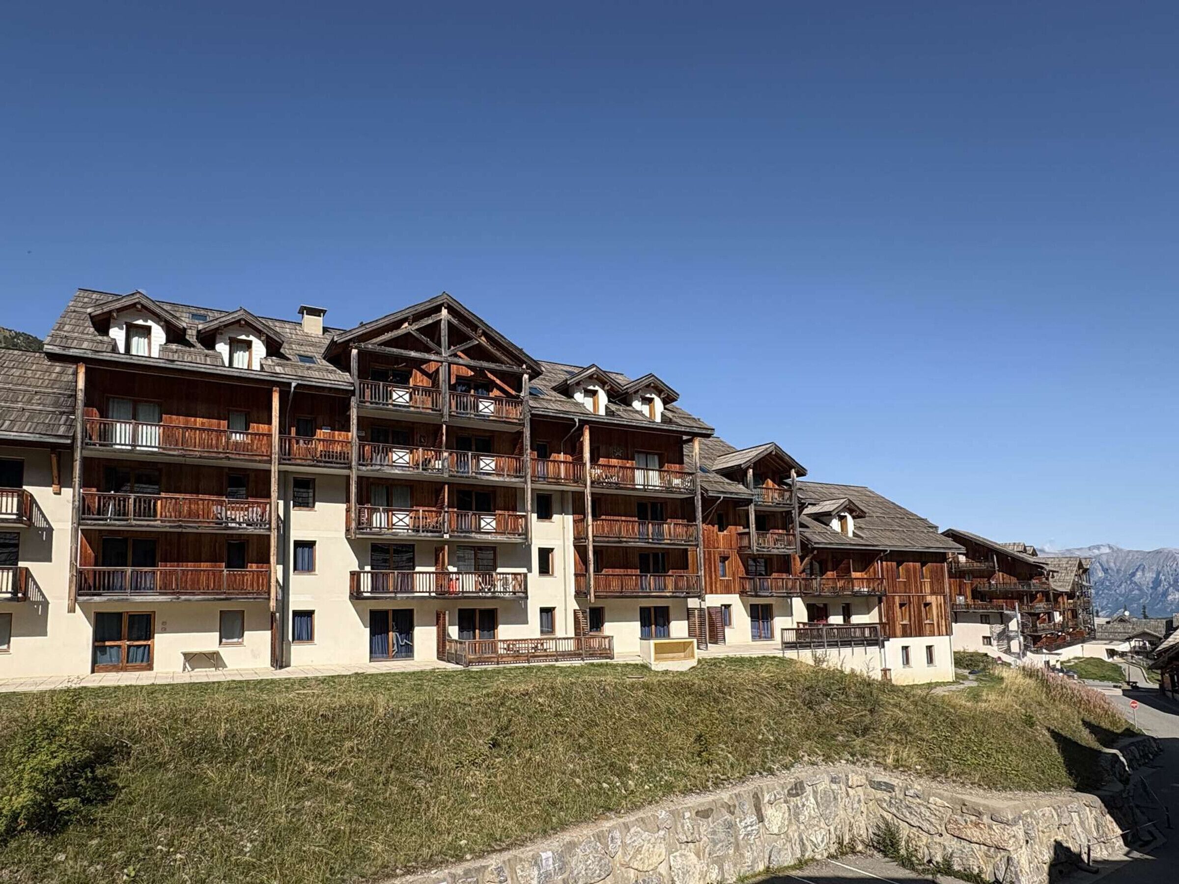 Un hotel de varios pisos con balcones, un muro de piedra y montañas al fondo.