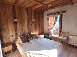 A wooden-paneled bedroom with a bed, two wall-mounted lamps, a window with curtains, and a chair. - Charming chalet with WiFi - 18 guests (Samoëns)