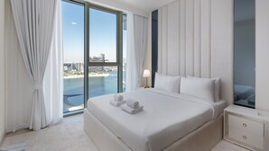 A hotel room with a large bed, a nightstand, a lamp, and a view of the city and water. - Modern Beachfront 3BR Apt | Near Marina & JBR (Dubai)