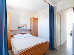 Ein Einzelbett mit Holzeinrahmung, ein Holzschrank, ein Regal mit einem Gemälde und eine Tür zur rechten Seite.