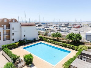 Ein Schwimmbecken, das von Grün umgeben ist, ein Gebäude mit Balkonen und ein Hafen mit Booten im Hintergrund.
