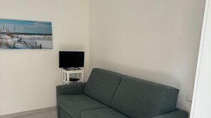 A room with a grey sofa, a flat-screen TV on a white stand, and a painting of a beach scene on the wall. - RAGGIO DI SOLE: STUDIO APARTMENT JUST A STONE'S THROW FROM THE BEACH (Castiglione della Pescaia)