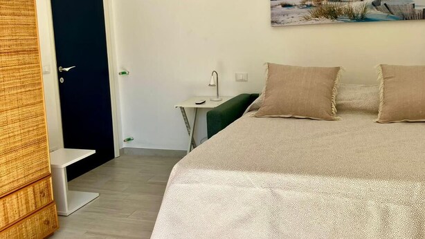 A bedroom with a bed, a nightstand, a lamp, a door, and an air conditioning unit. - RAGGIO DI SOLE: STUDIO APARTMENT JUST A STONE'S THROW FROM THE BEACH (Castiglione della Pescaia)