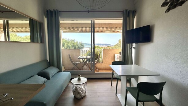 Et kompakt værelse med sofa, lille rundt bord, spisebord med stole, fjernsyn ophængt på væggen og en skydedør, der fører til en udendørs balkon.