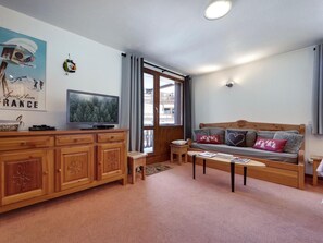 A cozy living room with a wooden sofa, a television on a wooden cabinet, and a window with a view of a building. - Ski-in/ski-out apartment, mountain view - 4 guests (Val-d'Isère)