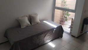 Quarto com cama, duas almofadas, janela com persianas e uma planta em vaso do lado de fora.