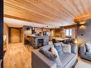 A modern interior with a wooden ceiling, a stone fireplace, a grey sofa, and a dining area with a large table and chairs. - Spacious apartment near slopes and shops - 8 guests (Val-d'Isère)