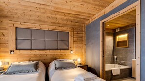 Ein Zimmer in einer Holzhütte mit zwei Betten, einem großen, gepolsterten Kopfteil, Nachttischlampen und einem Badezimmer mit Badewanne und Spiegel.