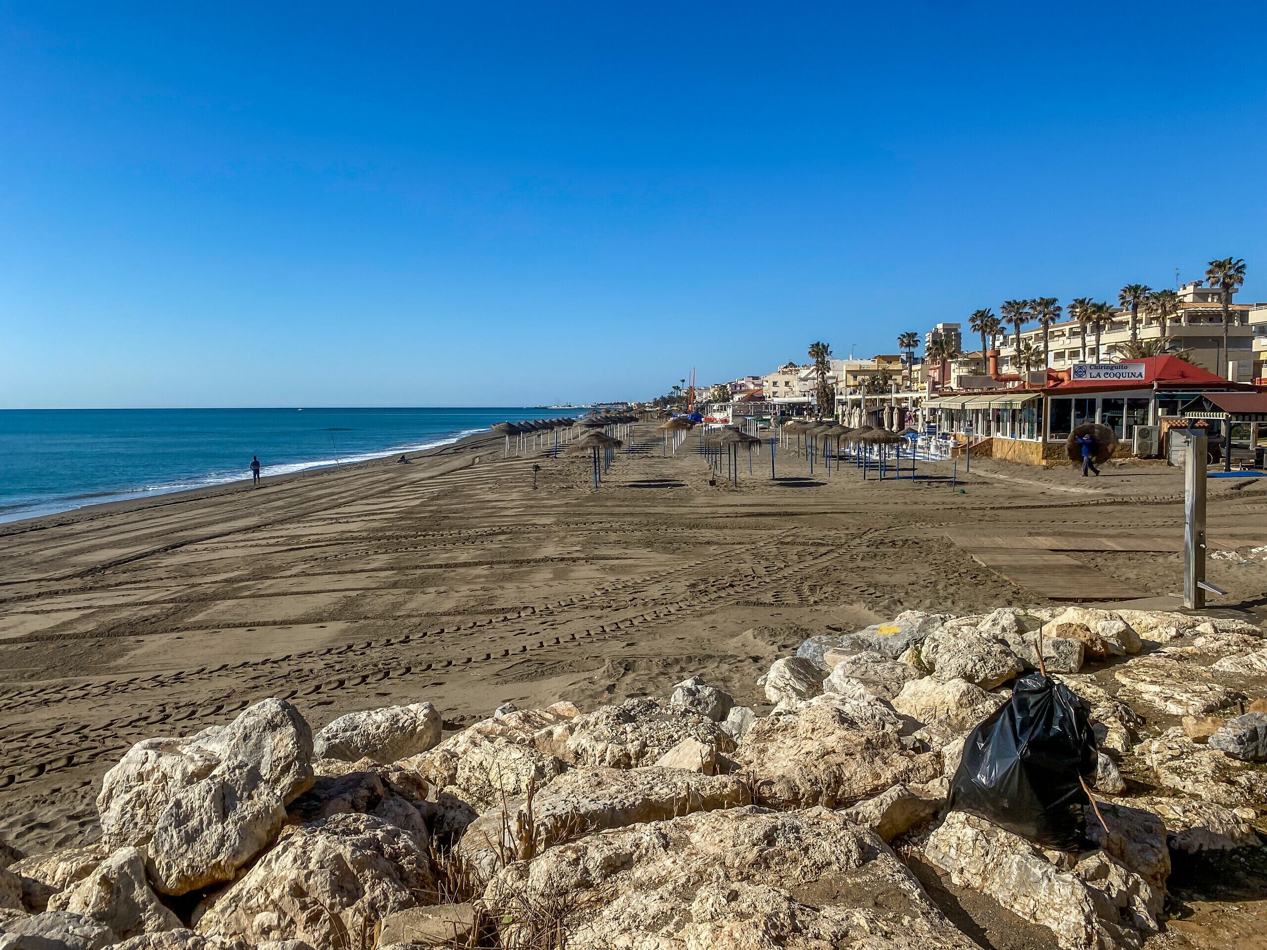 Ein Strand mit felsigem Vordergrund, einem Steg und Gebäuden mit Palmen im Hintergrund.