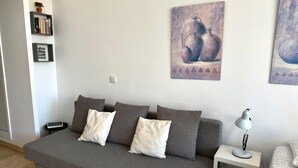 Harmaa sohva, jossa on valkoisia ja harmaita koristetyynyjä, valkoinen sivupöytä, jossa on lamppu, ja kaksi kehystettyä taideteosta seinällä.