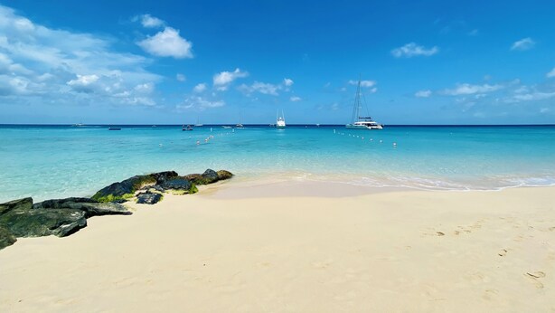 Ein Strand mit klarem türkisfarbenem Wasser, einem sandigen Ufer und mehreren Segelbooten in der Ferne.