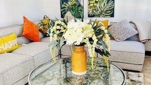 Ein Wohnzimmer mit Glastisch, einer Couch mit Kissen, einer Blumenvase und zwei Bildern an der Wand.
