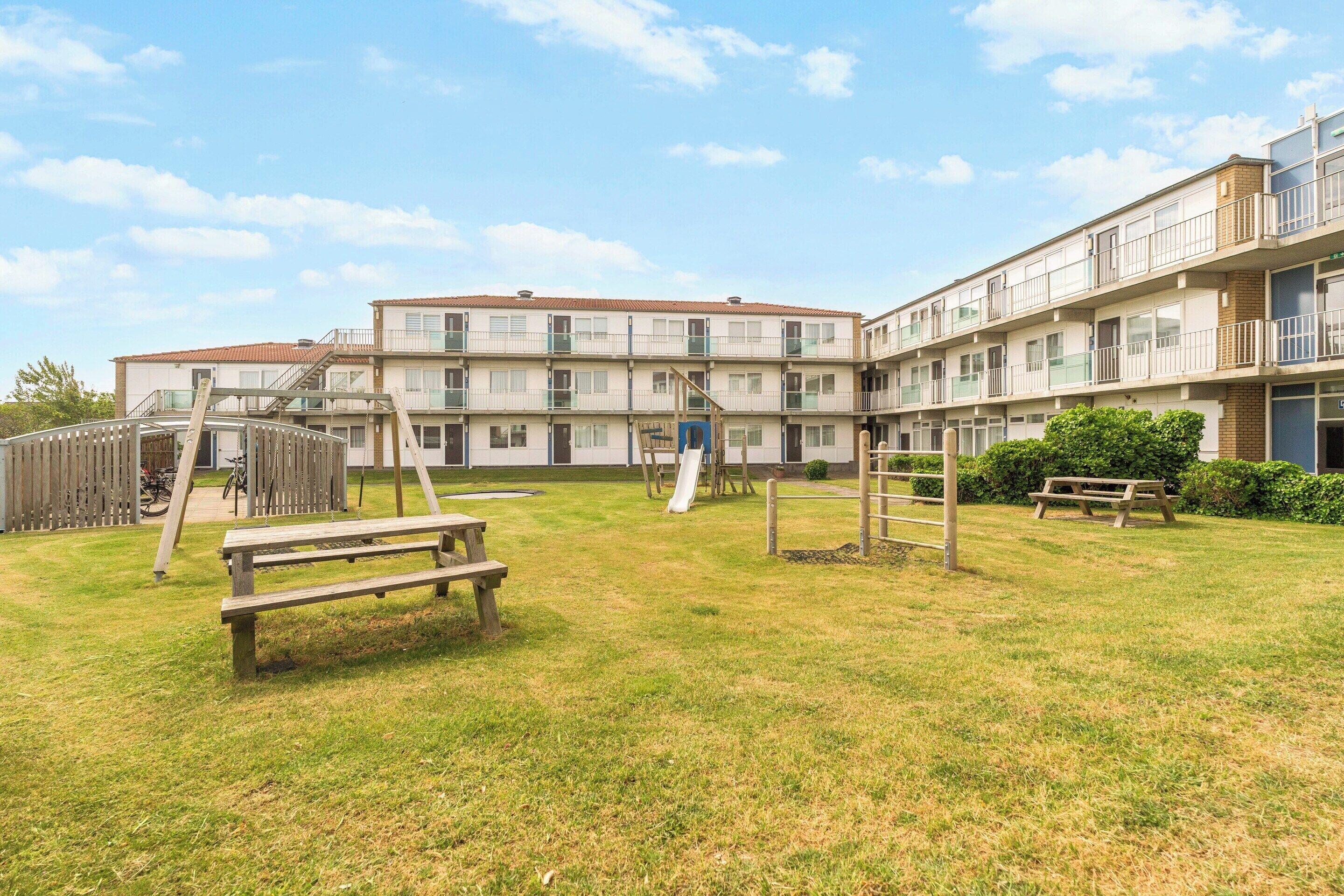 Um edifício residencial com vários andares, um parquinho, mesas de piquenique e um suporte para bicicletas.