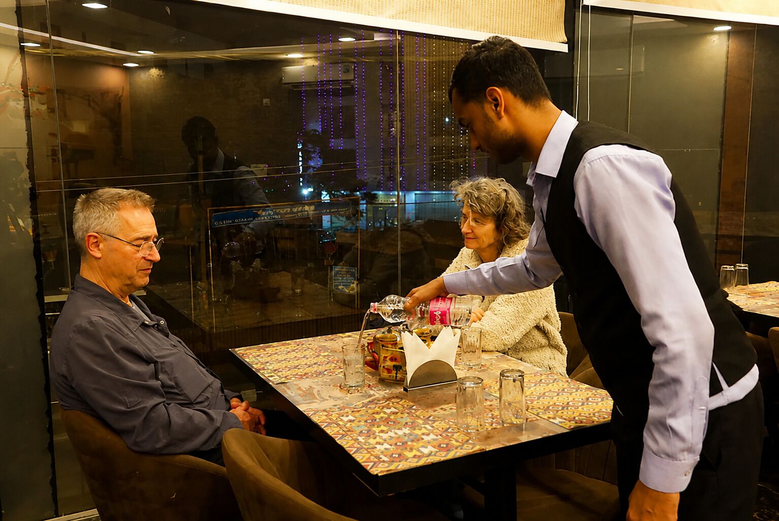 Ein Restaurantinnenraum mit einem Tisch für zwei Personen, einem Kellner, der einen Gast bedient, und einem weiteren Gast, der Platz nimmt.