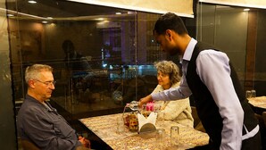 En restaurantindretning med et bord dækket til to personer, en tjener der tager sig af en gæst, og en anden gæst siddende.