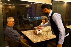 A restaurant interior with a table set for two, a waiter attending to a guest, and another guest seated.