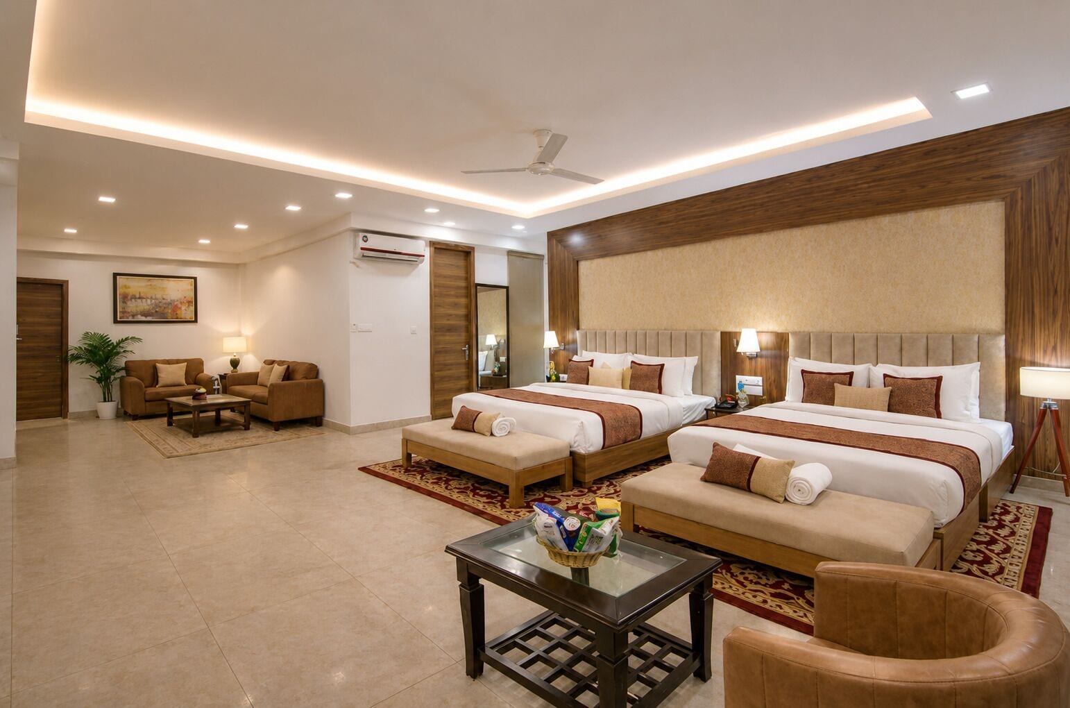 A hotel room with two beds, a sitting area with a sofa and armchairs, a small table with a basket, and a ceiling fan.