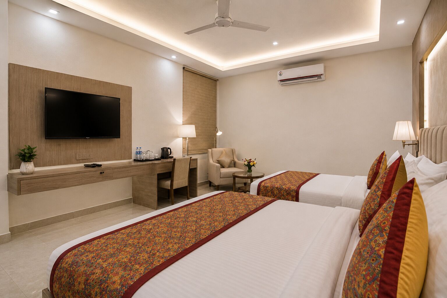 A hotel room with a flat-screen TV, a desk with a lamp, a chair, a sofa, two beds with patterned bedspreads, and an air conditioning unit.