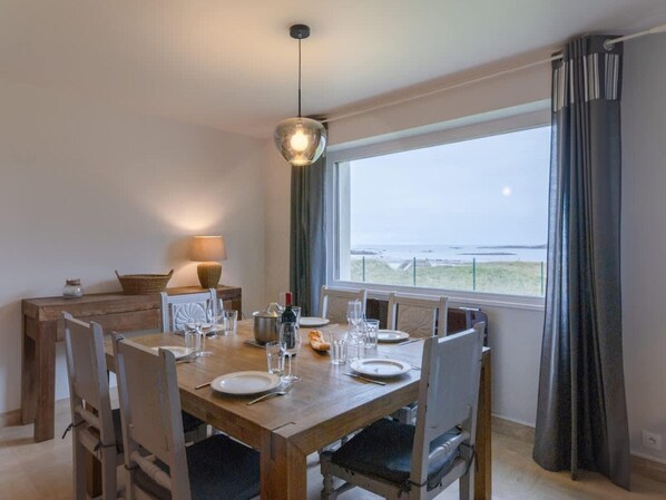 A dining area with a wooden table set for four, a hanging light fixture, and a view of the sea through large windows. - Magnificent Beach House for 6 (Ploudalmezeau)
