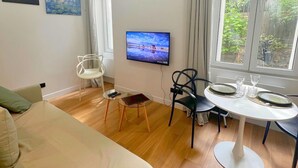 A modern room with a bed, a TV, a dining table with chairs, and a window with a view of greenery. - Comfortable & Peaceful Apartment, Latin Quarter - Newly Renovated (Paris)