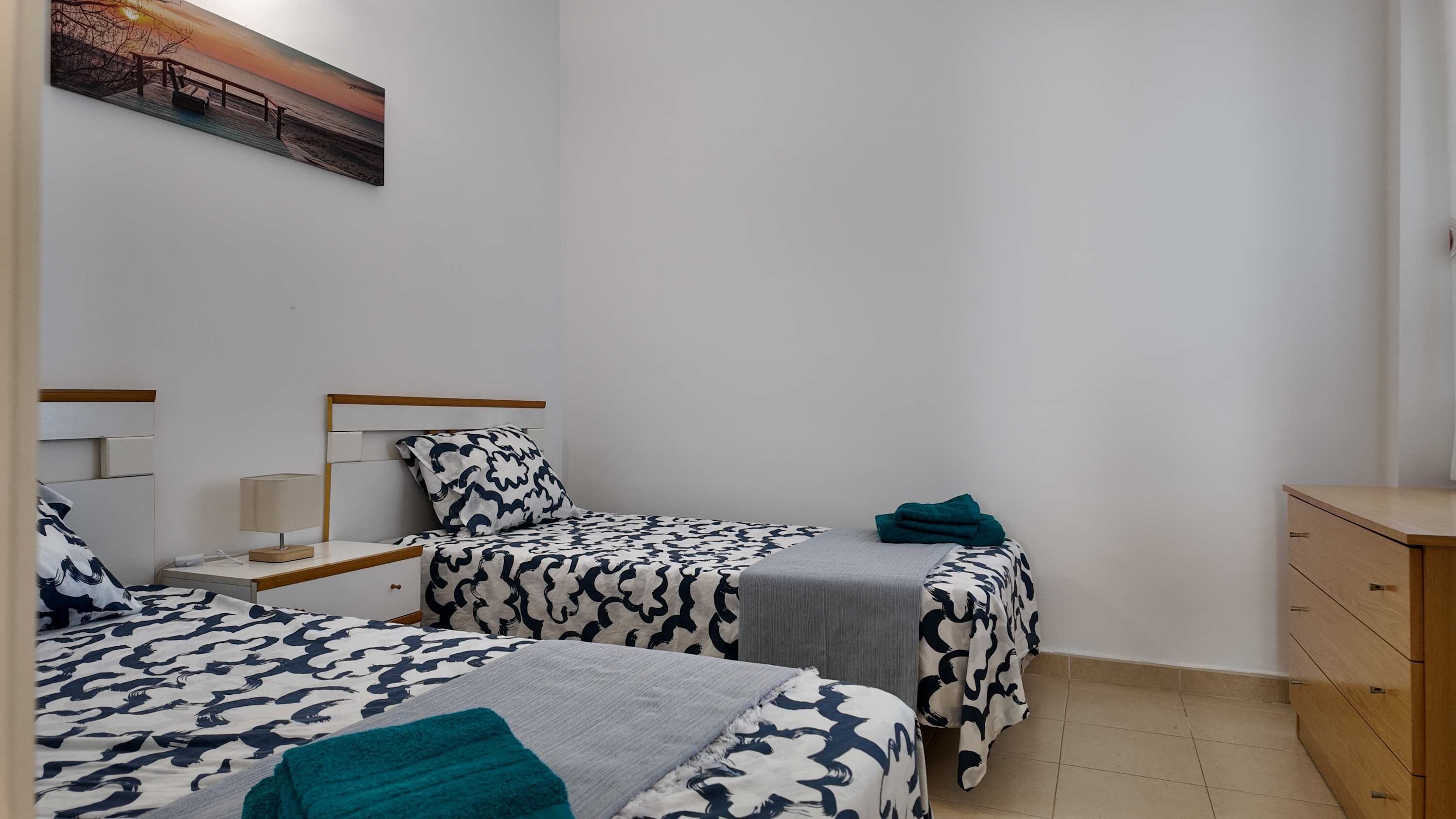 A bedroom with two beds, a nightstand, a lamp, a dresser, and a painting on the wall.