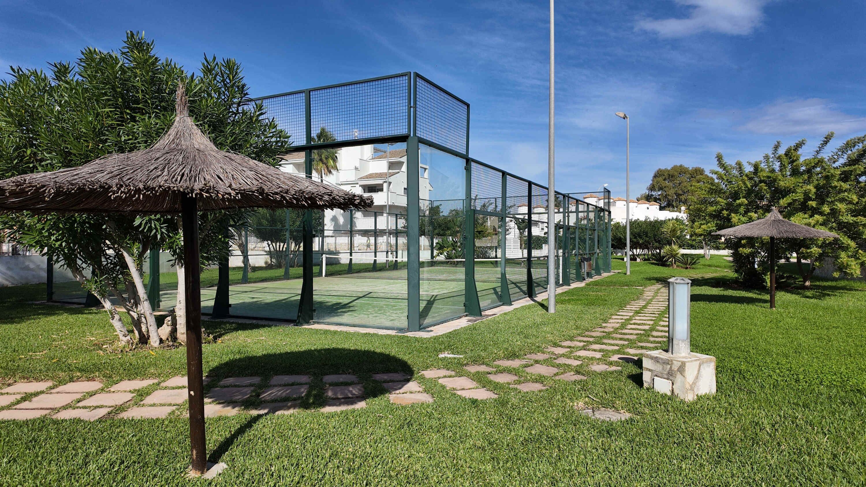 A tennis court with a net, surrounded by a green fence, and a thatched-roof pavilion.