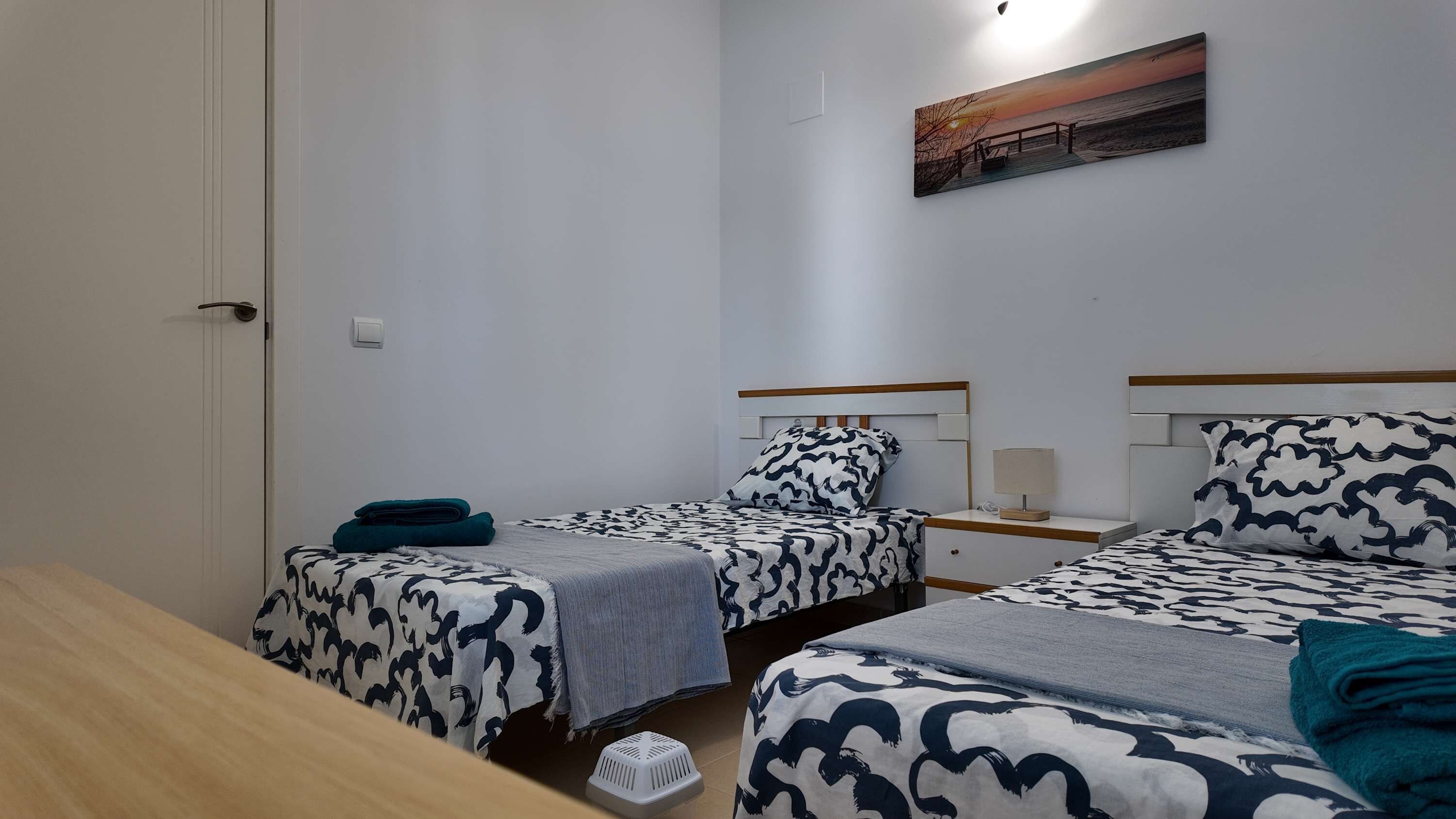 A room with two beds, a small table, a nightstand, a painting, and a door.