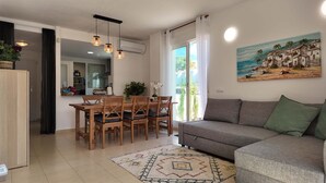A modern living room with a grey sofa, wooden dining table, and chairs. There is a painting of a beach scene on the wall. - Apartment in El Verger, 800 metres from the sea, with garden and communal pool (El vergel)