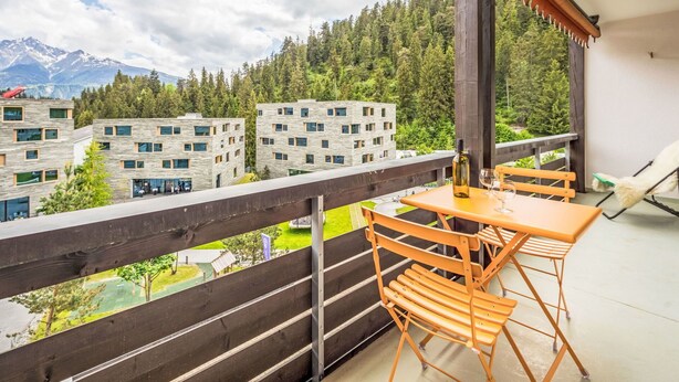 Un balcon avec une table et des chaises, donnant sur une zone boisée et sur des bâtiments avec une chaîne de montagnes en arrière-plan.
