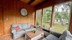 Una stanza accogliente con soffitto in legno, un divano grigio con cuscini rossi, un tavolino in legno e un'ampia finestra vista alberi e una casa.