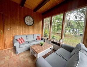 Ein gemütliches Zimmer mit Holzbalkendecke, einem grauen Sofa mit roten Kissen, einem Holztisch und einem großen Fenster mit Blick auf Bäume und ein Haus.