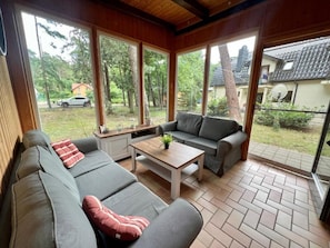 A sunlit room with a grey sofa, a wooden coffee table, and large windows offering a view of a residential area. - Holiday Home With Private Garden, Close to the sea (Rewal)