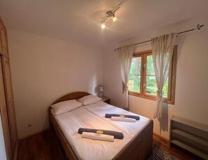 A bedroom with a wooden bed, two pillows, a nightstand with a lamp, a window with curtains, and a ceiling fan. - Holiday Home With Private Garden, Close to the sea (Rewal)