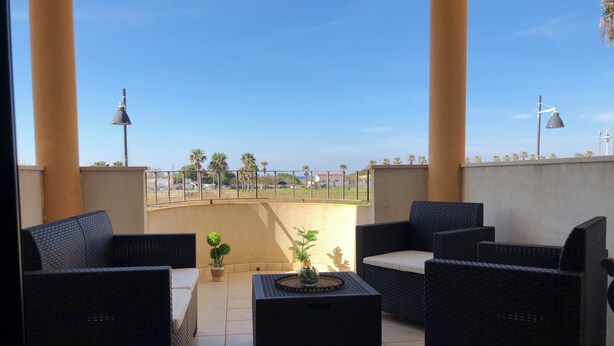 A balcony with wicker furniture, a potted plant, and a view of palm trees and a distant fence. - Townhouse just a stone's throw from the sea (Porto Cesareo)