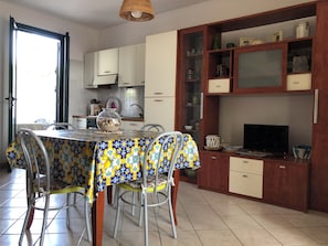 A dining area with a table set for four, a television on a wooden cabinet, and a kitchen in the background. - Townhouse just a stone's throw from the sea (Porto Cesareo)
