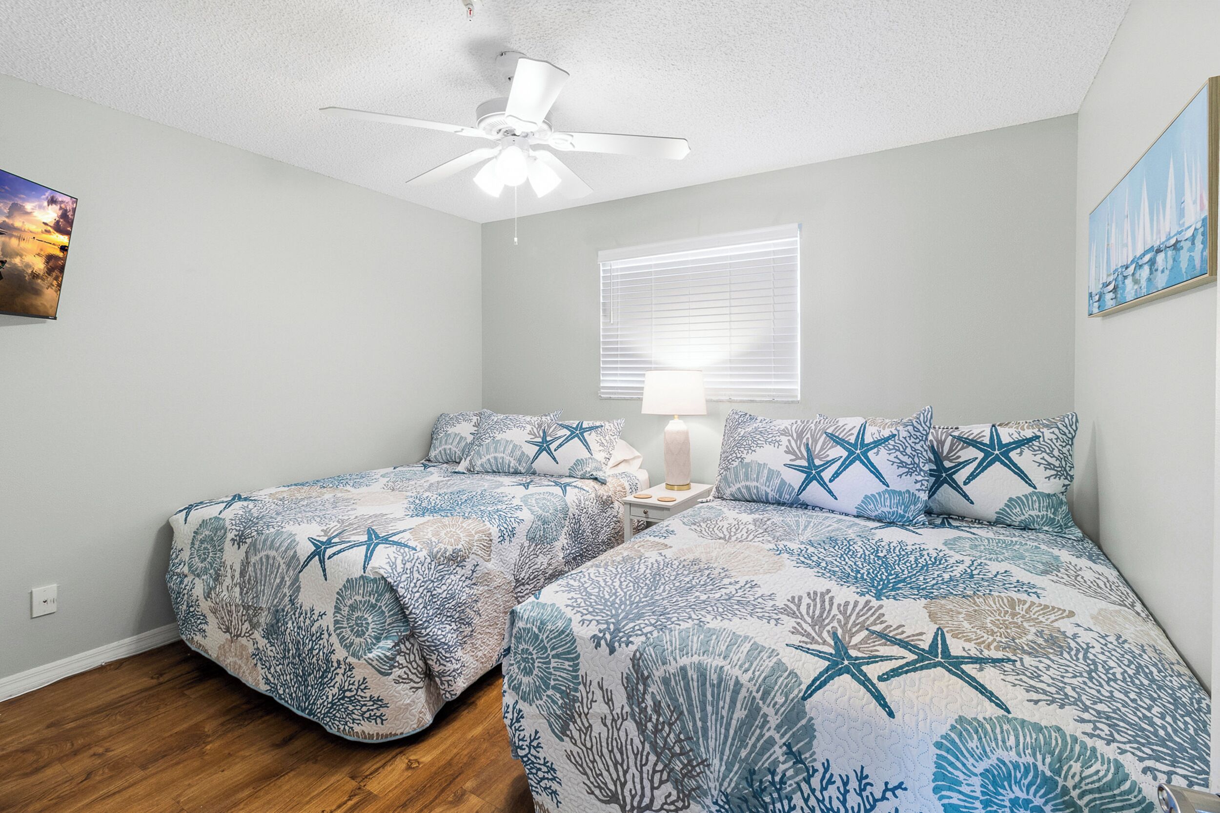 A bedroom with two beds, a ceiling fan, a window with blinds, and a wall-mounted painting.