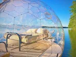 A transparent dome structure with a bed inside, a wooden floor, and a view of a lake.