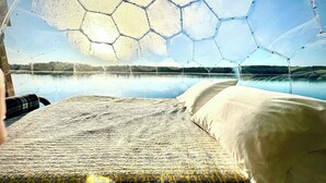Ein Bett mit einem gemusterten Bettbezug, einem weißen Kopfkissen und Blick auf einen See durch eine sechseckige Fensterkonstruktion.